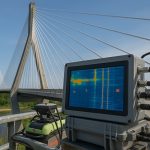 Bridge Health Monitoring Systems Transforming Safety and Efficiency A cable-stayed bridge with tall concrete pylons and steel cables, viewed from the side, featuring a monitoring device in the foreground displaying a heatmap of structural data.