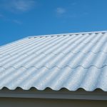White corrugated roof with visible condensation droplets under a clear blue sky, attached to a light-coloured building.
